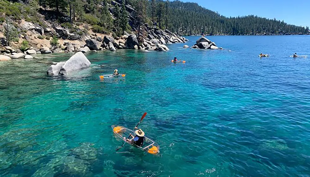 People are kayaking and paddleboarding on a clear blue lake surrounded by forested hills and rocky shores