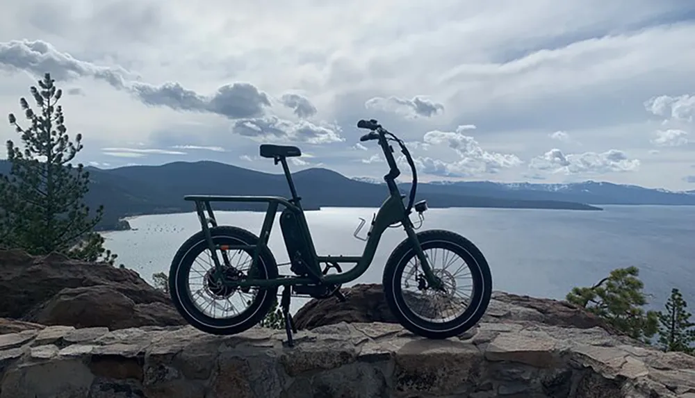 A bicycle stands silhouetted against an expansive view of a lake and mountains under a partly cloudy sky