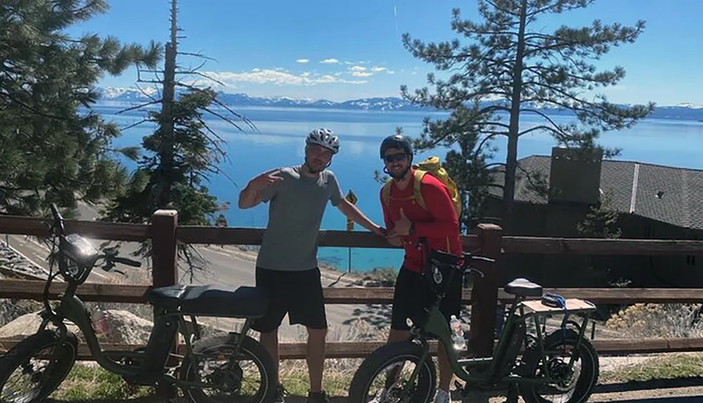 Two individuals in sporty attire are posing with bicycles against a scenic backdrop of a lake and mountains