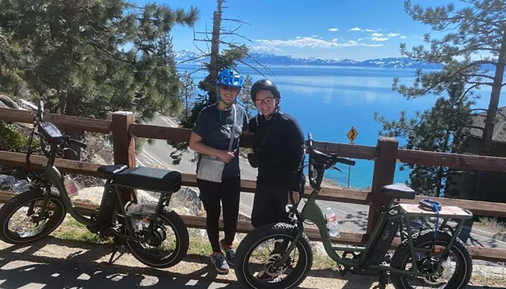 Two people with bike helmets are standing beside electric bikes with a scenic view of a lake and mountains in the background