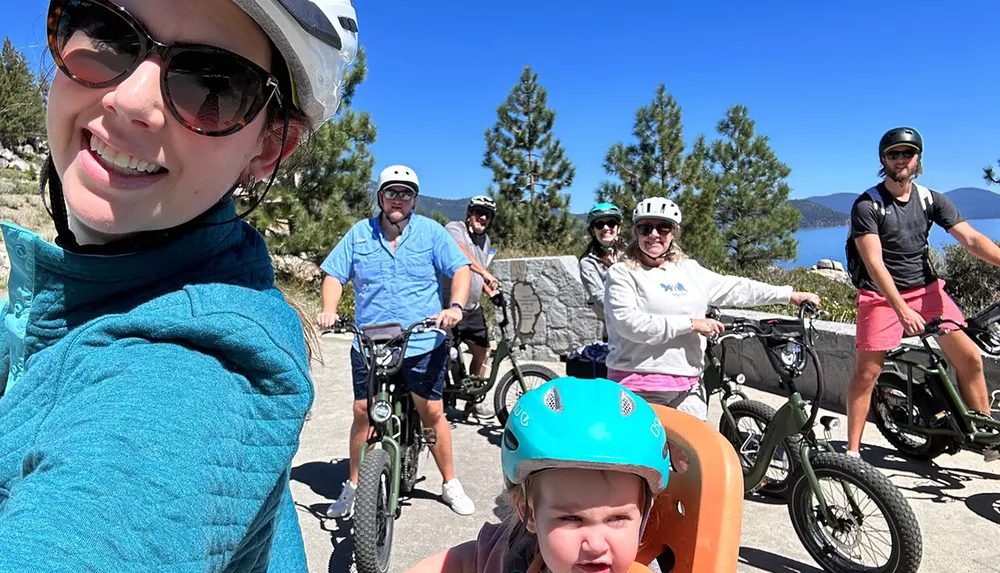 A group of people including a small child in the foreground are posing for a selfie while on a bike ride in a scenic location with clear skies and trees in the background