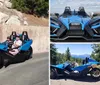 A person is driving a futuristic blue three-wheeled vehicle with a passenger beside them on a sunny day near a concrete wall displaying a No Parking Any Time sign