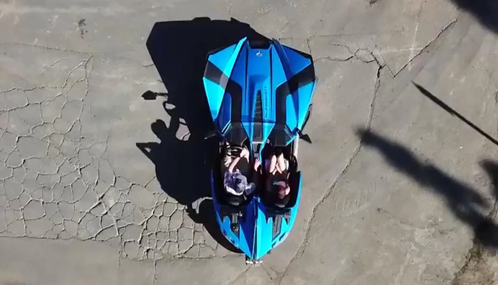 The image shows an overhead view of a vibrant blue sports car with two people inside casting a long shadow on a cracked concrete surface