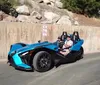 A person is driving a futuristic blue three-wheeled vehicle with a passenger beside them on a sunny day near a concrete wall displaying a No Parking Any Time sign