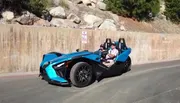 A person is driving a futuristic blue three-wheeled vehicle with a passenger beside them on a sunny day near a concrete wall displaying a 