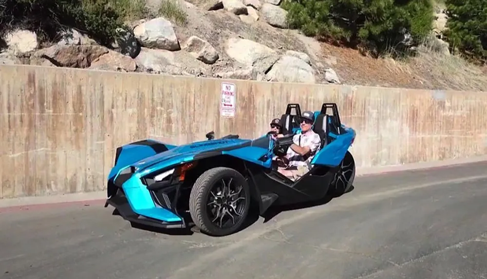 A person is driving a futuristic blue three-wheeled vehicle with a passenger beside them on a sunny day near a concrete wall displaying a No Parking Any Time sign