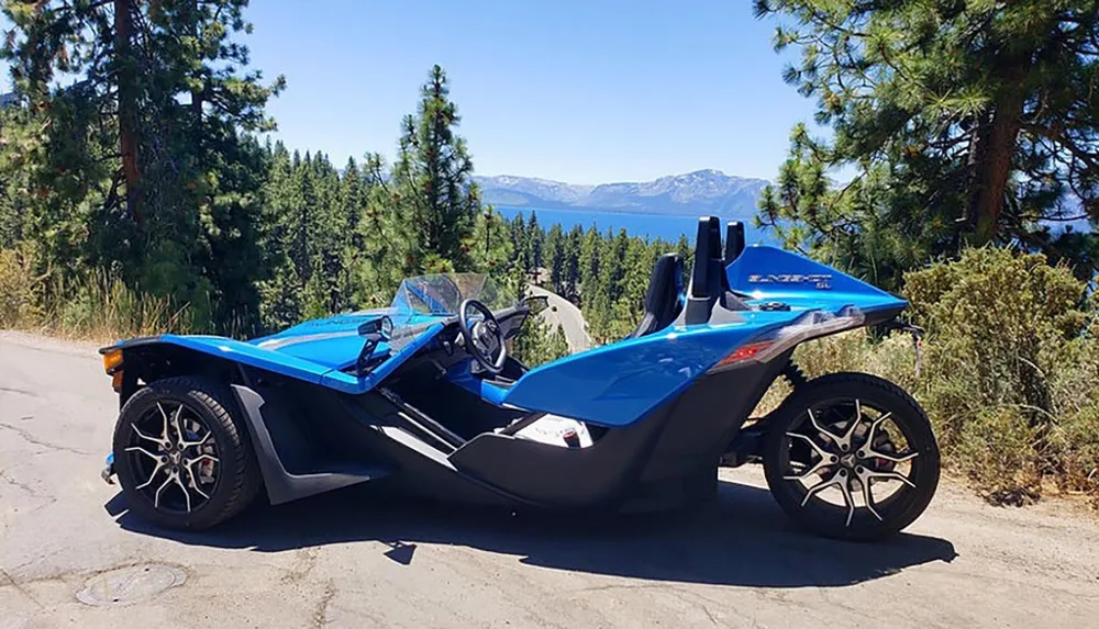 A blue three-wheeled vehicle is parked on the side of a road with a scenic view of a forest and a lake in the background