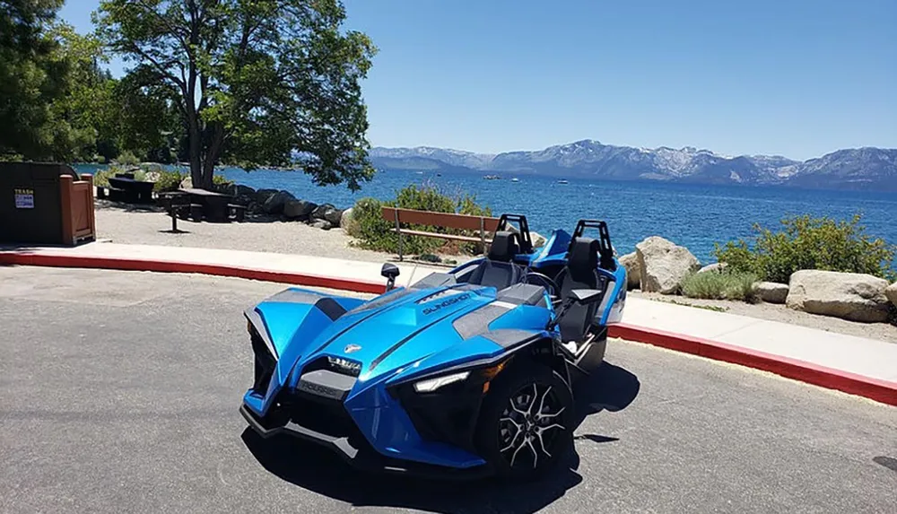A blue three-wheeled vehicle possibly a Polaris Slingshot is parked by the shore of a lake with mountains in the distance