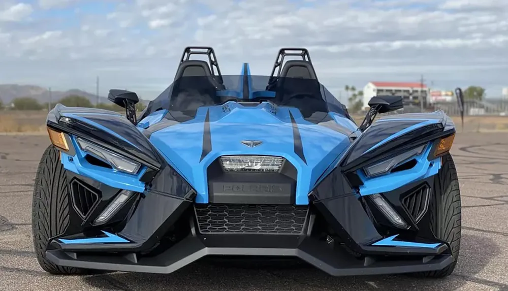 The image showcases a blue and black Polaris Slingshot a three-wheeled motor vehicle parked on an asphalt surface with overcast skies and distant scenery in the background
