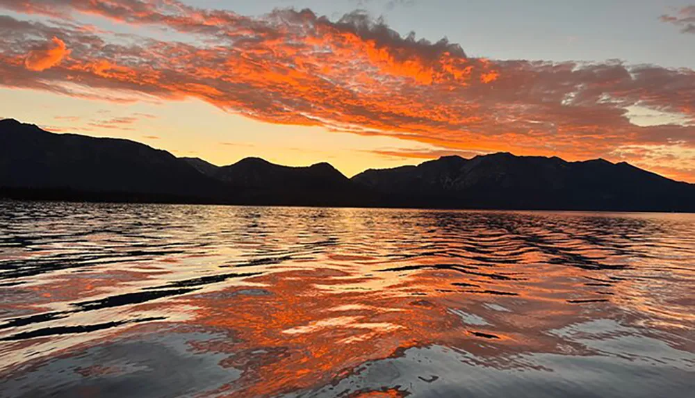 A fiery sunset paints the sky and reflects on the rippled surface of a lake against a backdrop of dark mountains
