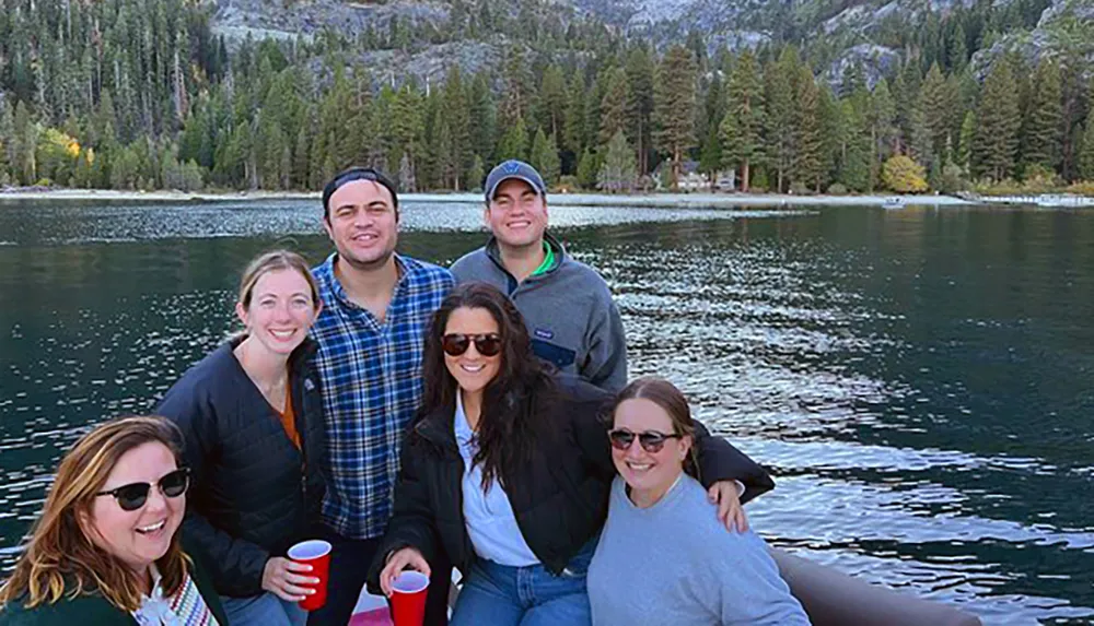 A group of six smiling people pose together for a photo with a backdrop of a serene lake and forested mountains