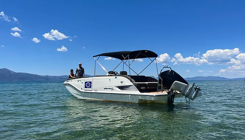 Two people stand aboard a moored boat with a canopy on a serene clear blue lake under a sunny sky dotted with a few clouds