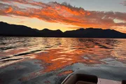 A vibrant sunset paints the sky with shades of orange and red above a serene mountain-lined lake, as seen from the edge of a boat.
