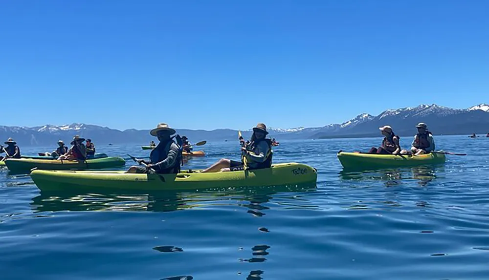 A group of people are kayaking in calm blue waters with a backdrop of distant snow-capped mountains under a clear sky
