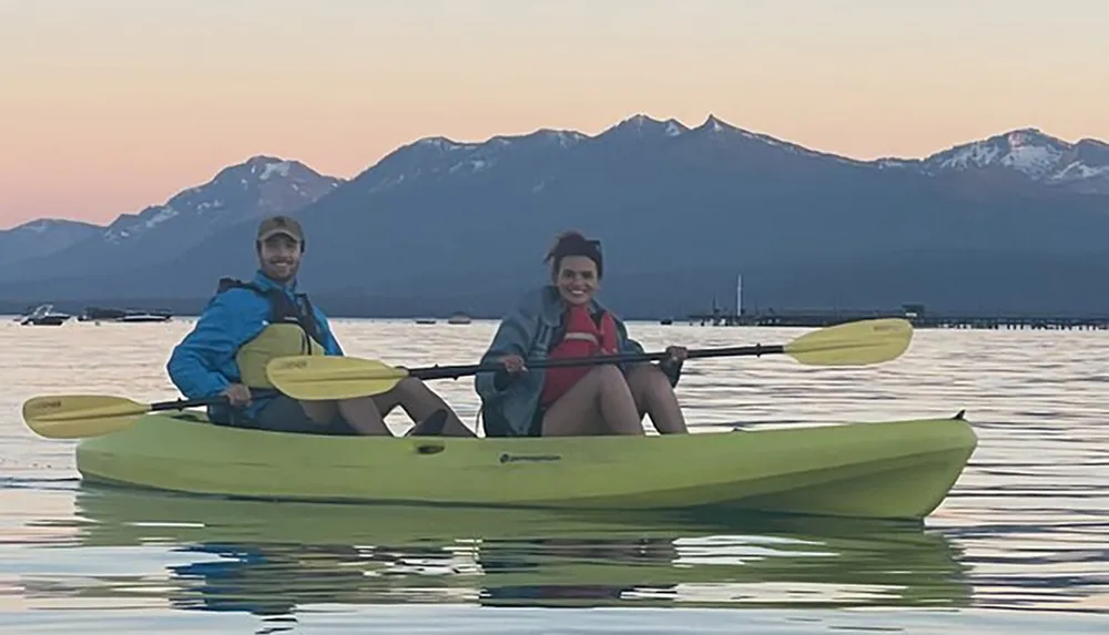 Two smiling people are kayaking on calm water with a stunning backdrop of mountains at sunset