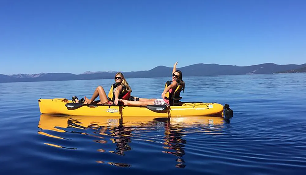Two people are enjoying a sunny day kayaking on a calm blue lake with mountains in the background
