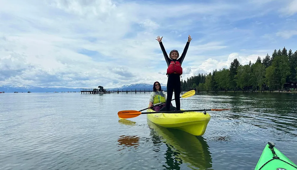 A child stands triumphantly atop a yellow kayak with a seated adult paddling behind in a serene lake surrounded by trees and mountains under a partly cloudy sky