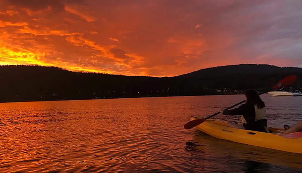 A person kayaks on a calm lake under a vibrant orange sunset sky