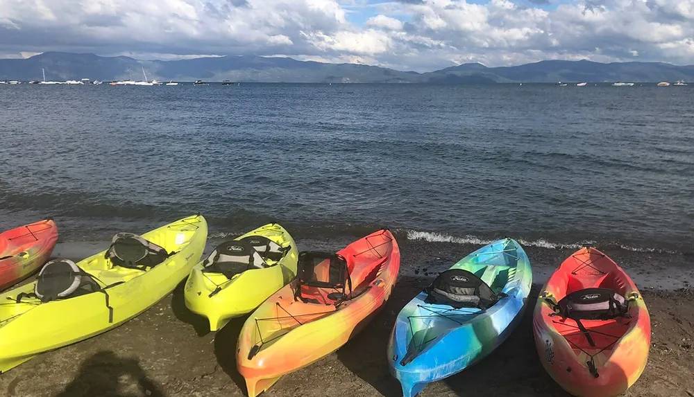 A line of colorful kayaks is arranged on a sandy shore with a view of a tranquil sea and distant mountains