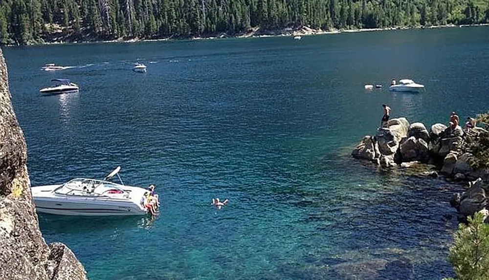 Several people enjoy a sunny day near the clear blue waters of a lake with boats floating nearby and a rocky shoreline in the foreground