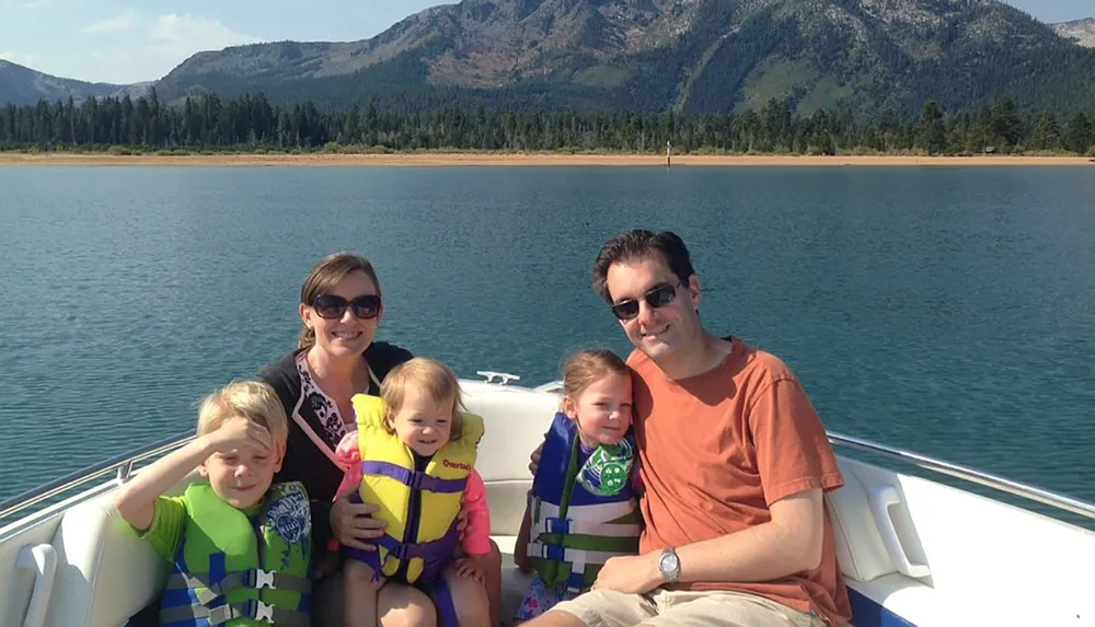 A family of two adults and three children is enjoying a sunny day on a boat with a scenic mountainous backdrop
