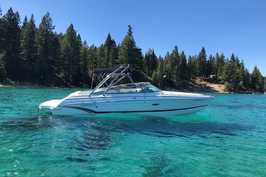 A white and blue motorboat is anchored in clear turquoise waters near a pine-tree-lined shore under a blue sky.