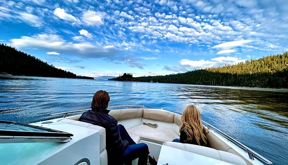 Two people are enjoying a boat ride on a serene lake surrounded by lush forests under a sky dotted with white clouds