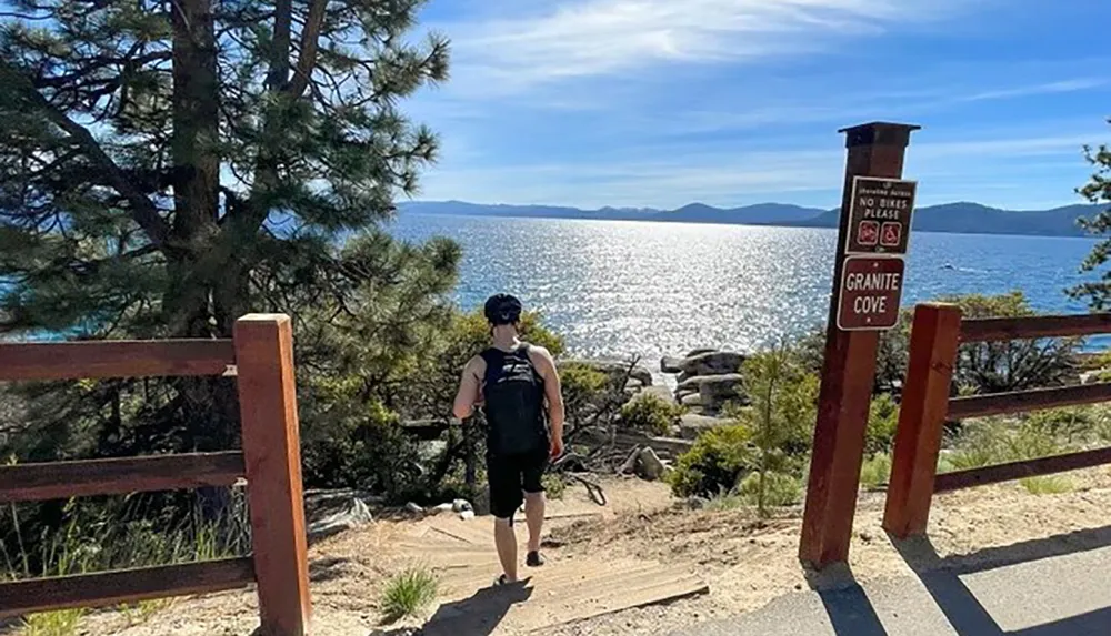 A person stands at the entrance of a trail overlooking a scenic view of a large lake backed by distant mountains