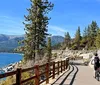 A group of people are posing with their bicycles on a sunny day near a picturesque lake surrounded by mountains and trees