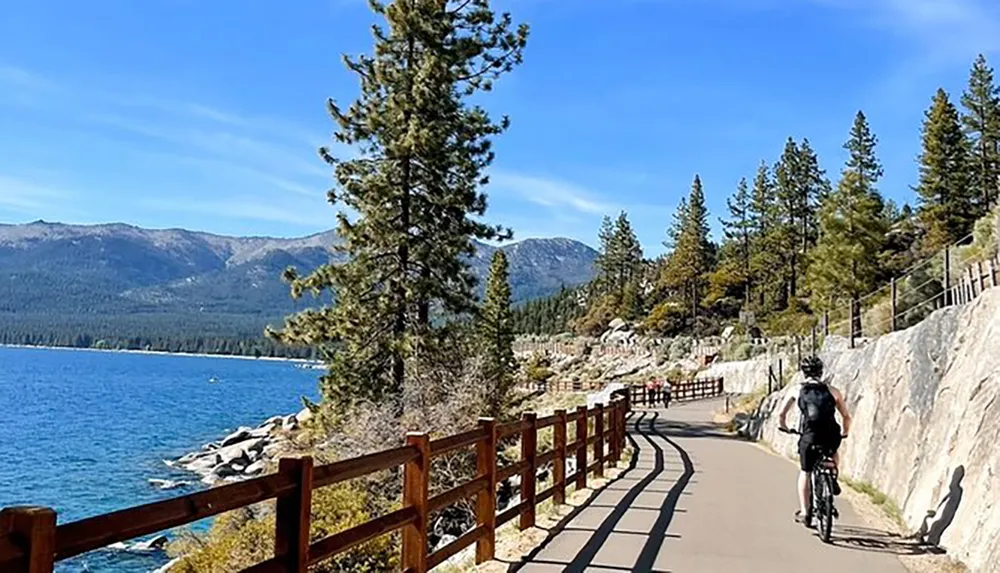 A person is biking along a scenic lakeside path flanked by a wooden fence and towering pine trees against a backdrop of mountains