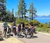 A group of people are posing with their bicycles on a sunny day near a picturesque lake surrounded by mountains and trees