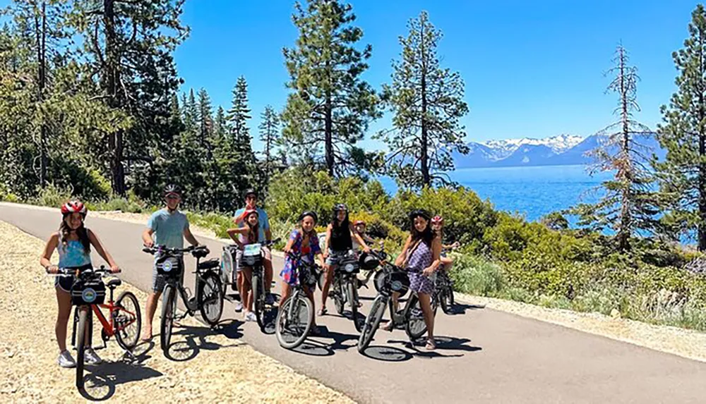 A group of people are posing with their bicycles on a sunny day near a picturesque lake surrounded by mountains and trees