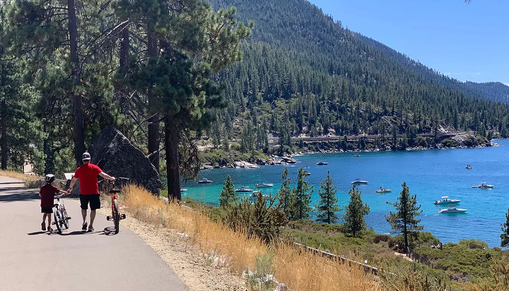 An adult and a child are walking with bicycles along a scenic lakeside path with a clear blue lake and pine-covered hills in the background