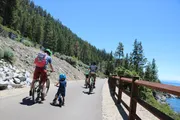 A group of four cyclists, one being a young child, are riding on a paved trail with a scenic forest landscape and a lake in the background under a clear blue sky.
