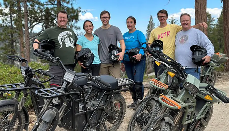 A group of six individuals stands smiling behind a line of electric bikes in an outdoor setting, holding their helmets and displaying a sense of camaraderie.