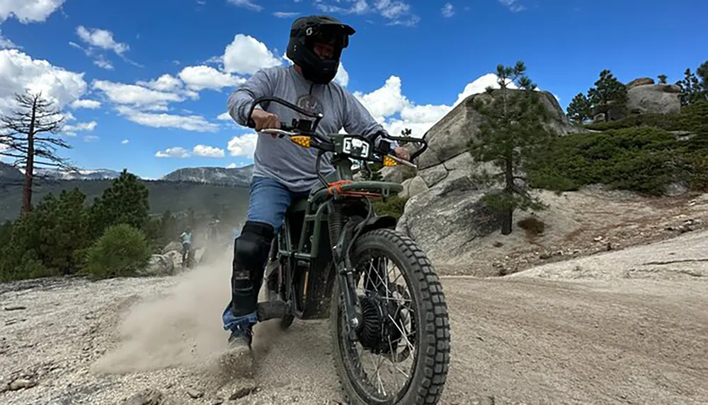A motorcyclist kicks up dust while riding on a rugged trail through a mountainous landscape