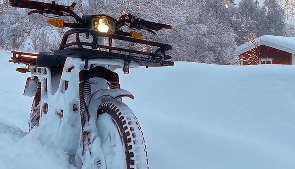 The image displays a snow-covered motorcycle with its headlight illuminated set against a snowy landscape with a cabin in the background