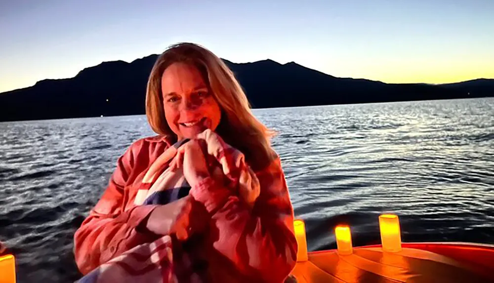 A person is smiling while sitting by a body of water with a mountain silhouette in the background and candles illuminating the scene at dusk