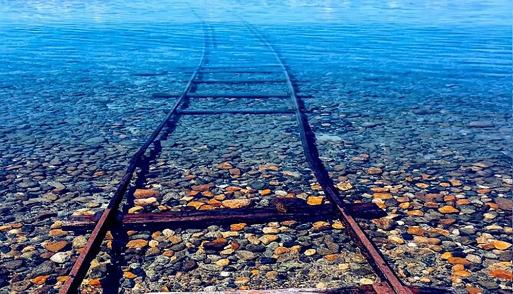 An old rusted railroad track leads into a clear blue body of water creating a tranquil yet thought-provoking scene