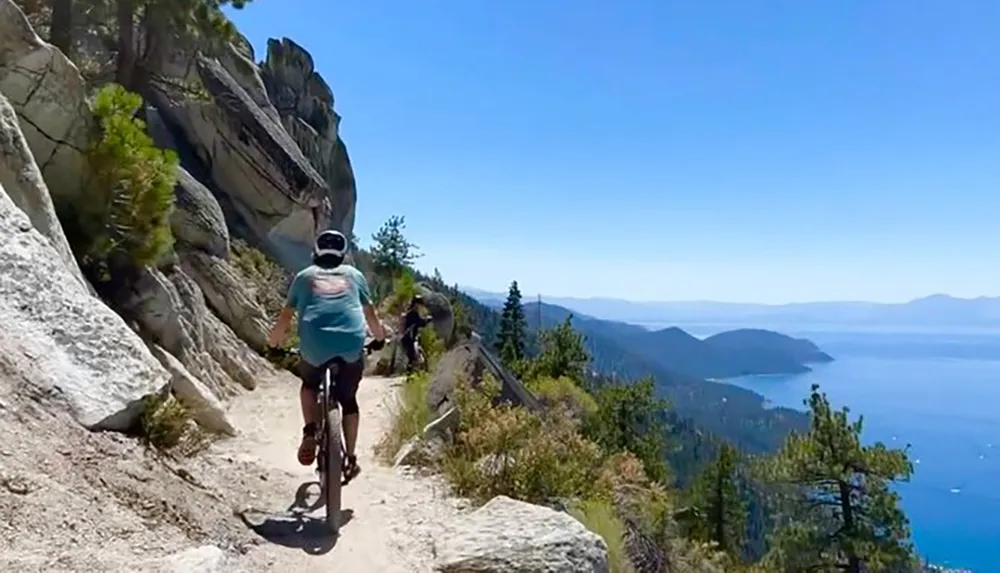 A person is mountain biking on a narrow trail along a steep mountainside with a breathtaking view of a blue lake in the distance