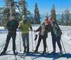 Four people are smiling for the camera while standing on a snowy slope with cross-country skis against a backdrop of snow-covered trees and a blue lake in the distance