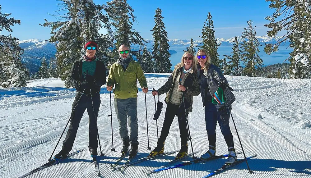 Four people are smiling for the camera while standing on a snowy slope with cross-country skis against a backdrop of snow-covered trees and a blue lake in the distance