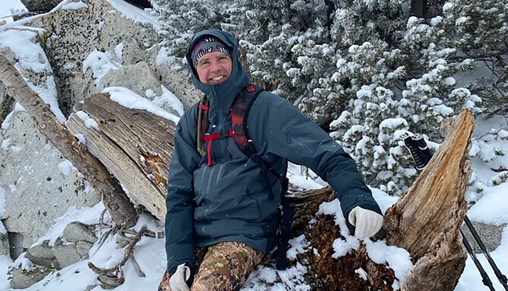 A person is sitting on a fallen tree trunk surrounded by snow-covered foliage smiling towards the camera dressed in winter attire suitable for hiking or outdoor activity
