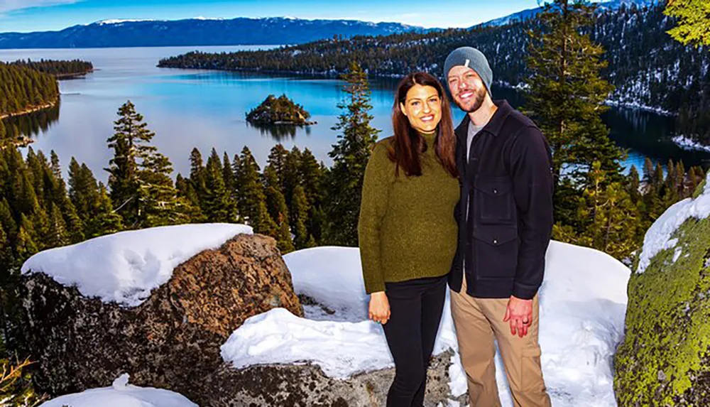 A smiling couple poses in winter clothing against a scenic backdrop featuring a snow-capped forest and a calm blue lake