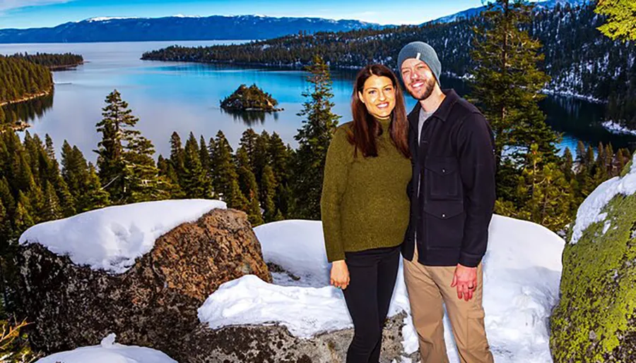 A smiling couple poses in winter clothing against a scenic backdrop featuring a snow-capped forest and a calm blue lake.