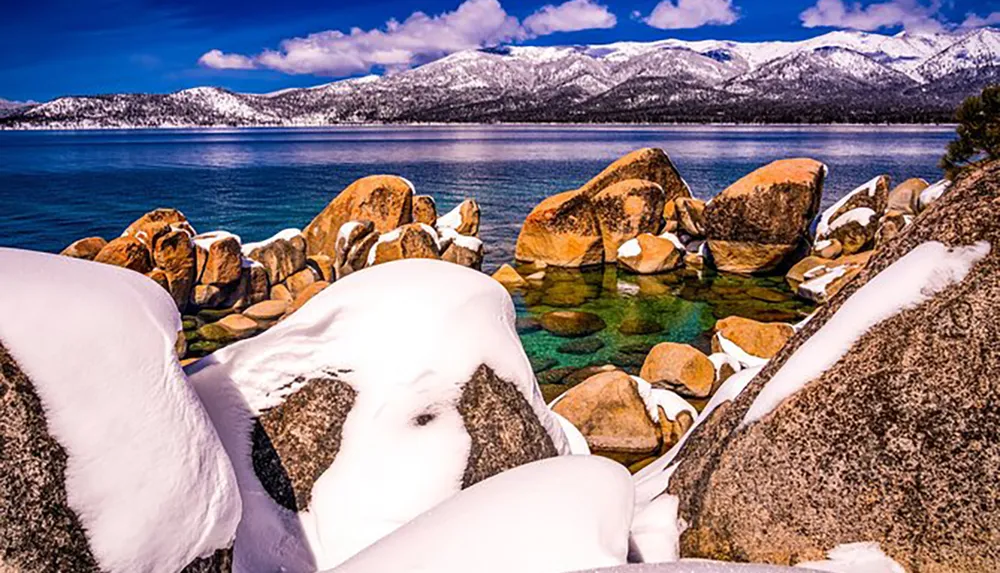 The image shows a tranquil winter scene with snow-covered boulders on the shore of a clear lake with a backdrop of snow-dusted mountains and a partly cloudy sky