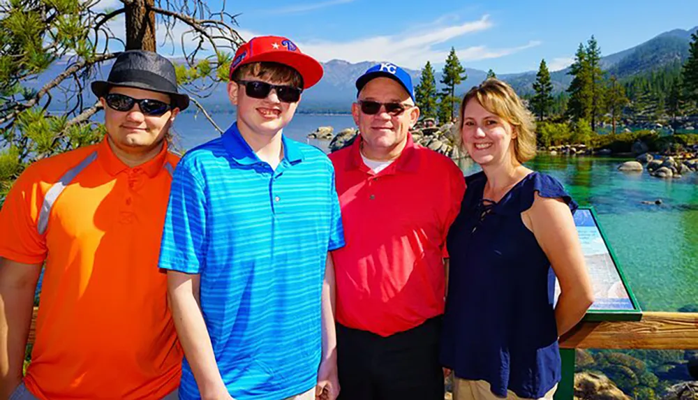 Four people are smiling for a group photo in front of a serene lake with clear waters and a pine forested mountainous backdrop