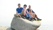 The image shows four individuals of varying ages sitting closely together atop a large rock with a seemingly calm body of water and a hazy sky in the background.