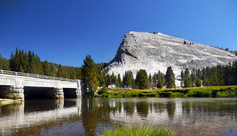 The image shows a serene landscape with a prominent granite dome beside a reflective river and a stone bridge with clear blue skies and trees around suggesting a natural park setting