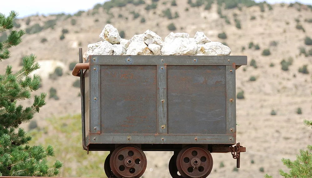 An old rusted mining cart is filled with large rocks set against a backdrop of hilly terrain and sparse vegetation